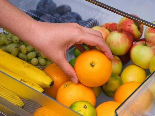 果物でいっぱいの冷蔵庫の引き出しからオレンジを取り出している